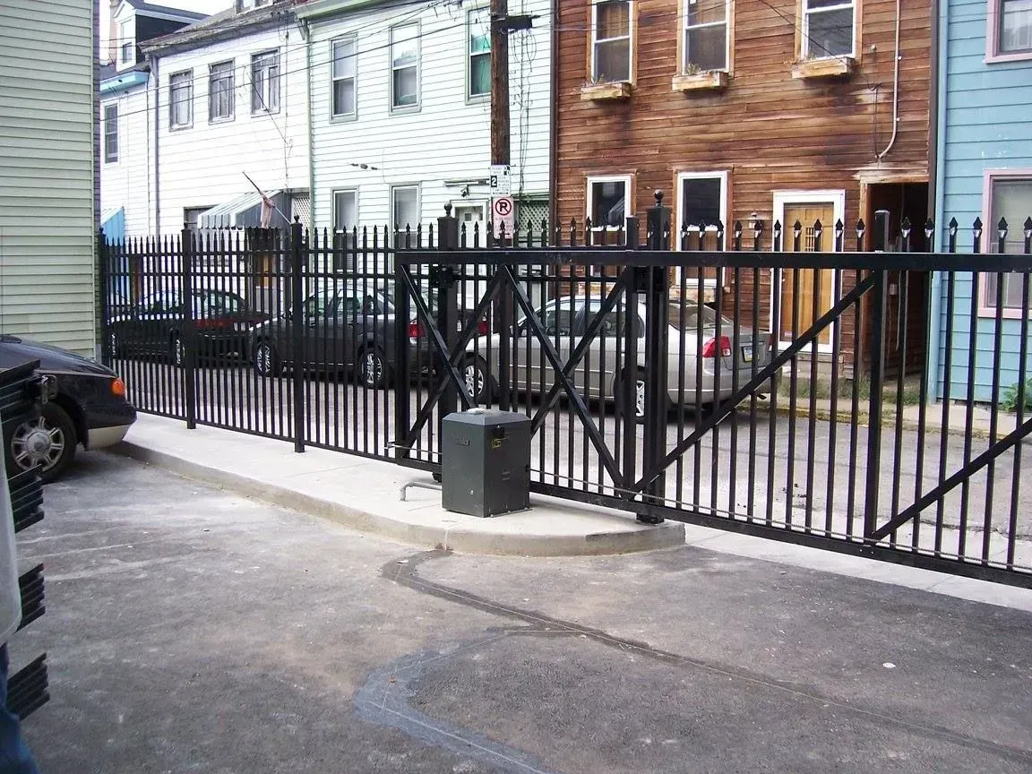 Black metal fence and gate across an alleyway with cars parked beyond. Buildings are visible in the background.