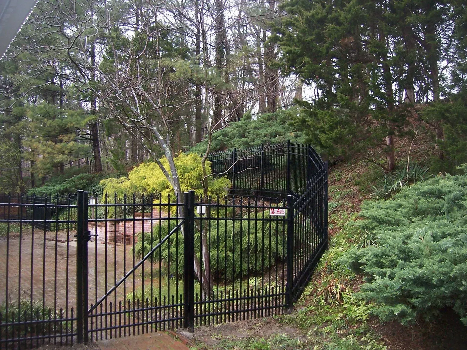 Black metal fence enclosing a garden area with lush greenery and trees in the background.