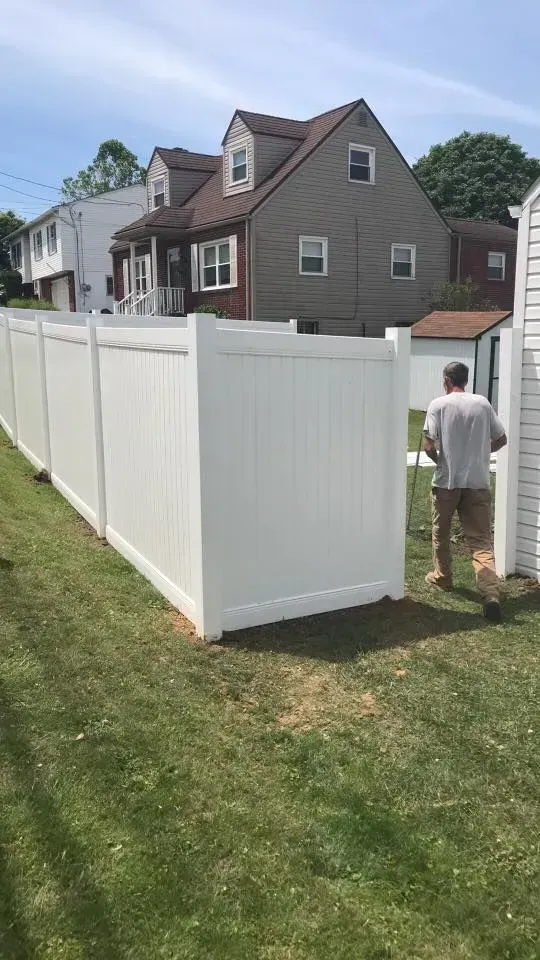 White vinyl fence in a grassy yard, man walking through a gate near a beige house.