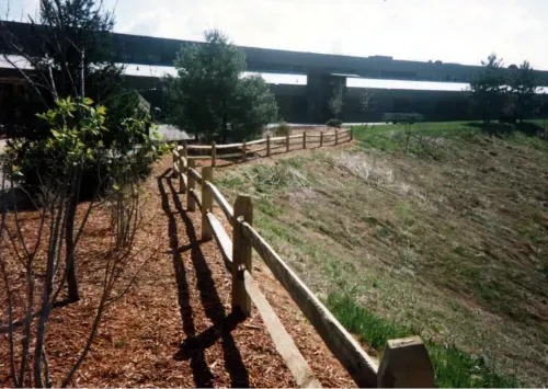 Wooden fence borders a sloped grassy hill with a building in the background. Brown mulch and small trees are also visible.