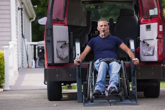 Man in wheelchair exiting a red accessible van using a ramp. He is outside on a driveway.