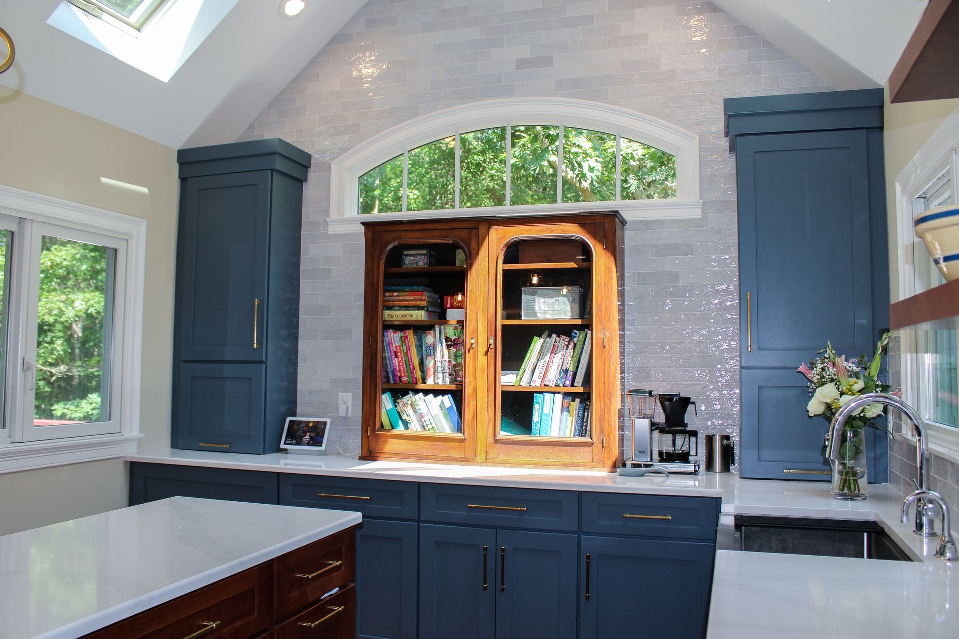 Kitchen with blue cabinets, a wooden bookcase, and an arched window overlooking trees.