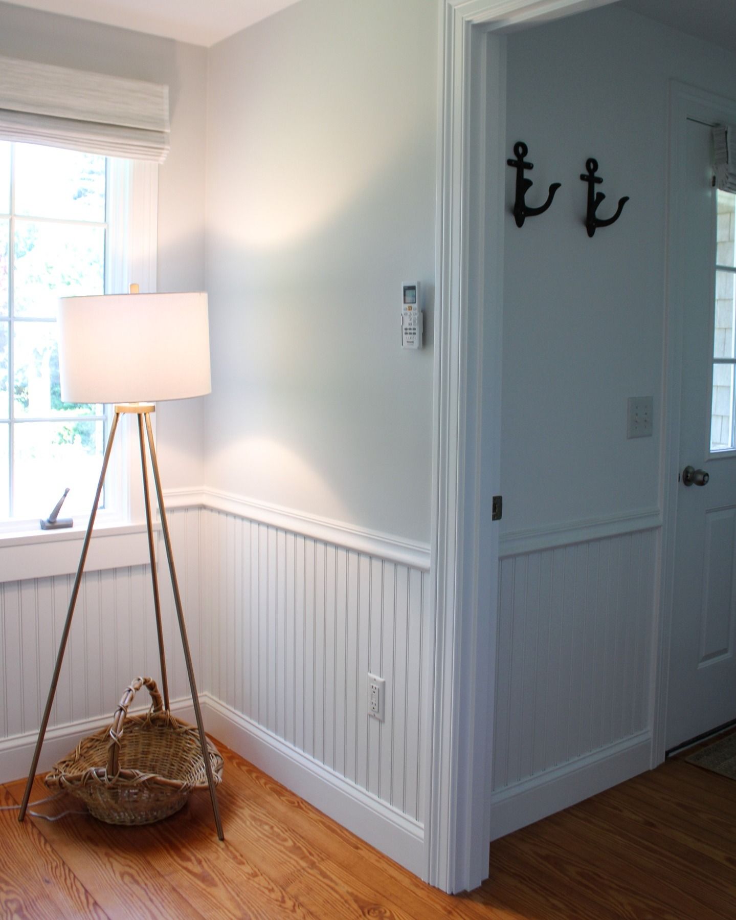 A coastal-style interior with a tripod lamp, basket, and white wainscoting. An open doorway with anchor hooks is visible.