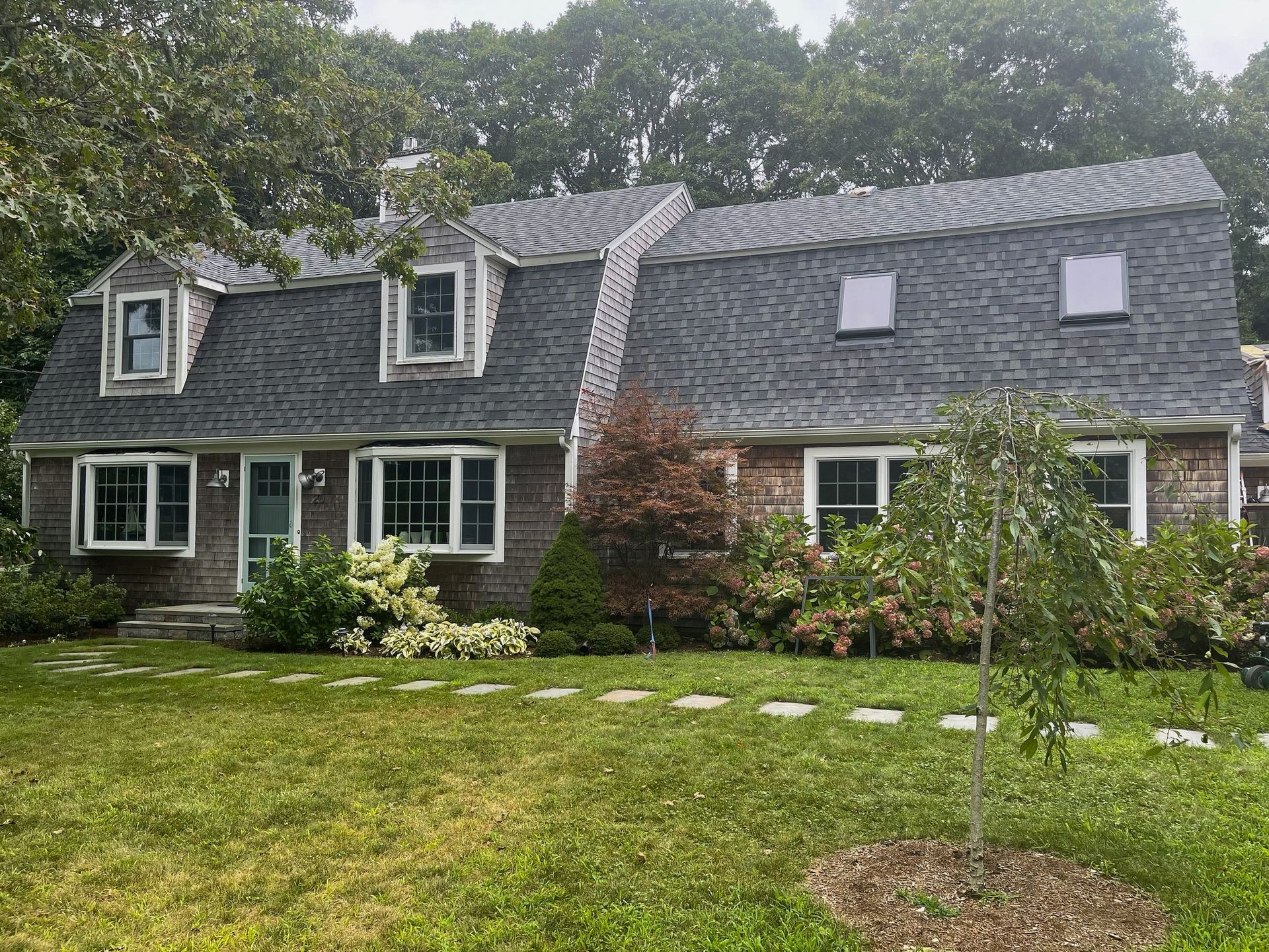 House with cedar shake siding, gray roof, dormers, and a green lawn.