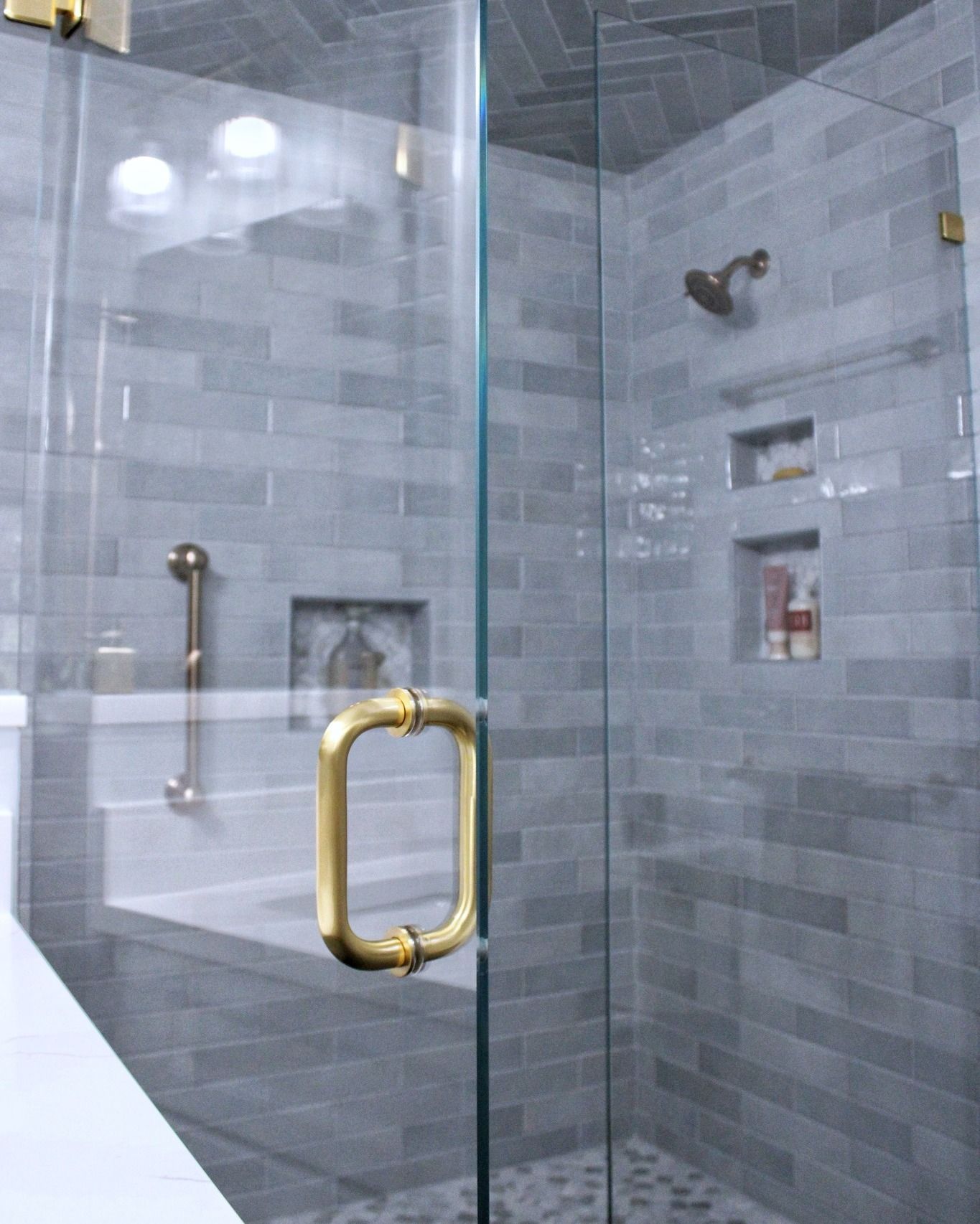 Gray tiled shower with glass door and gold hardware. Features a built-in bench and recessed shelves.