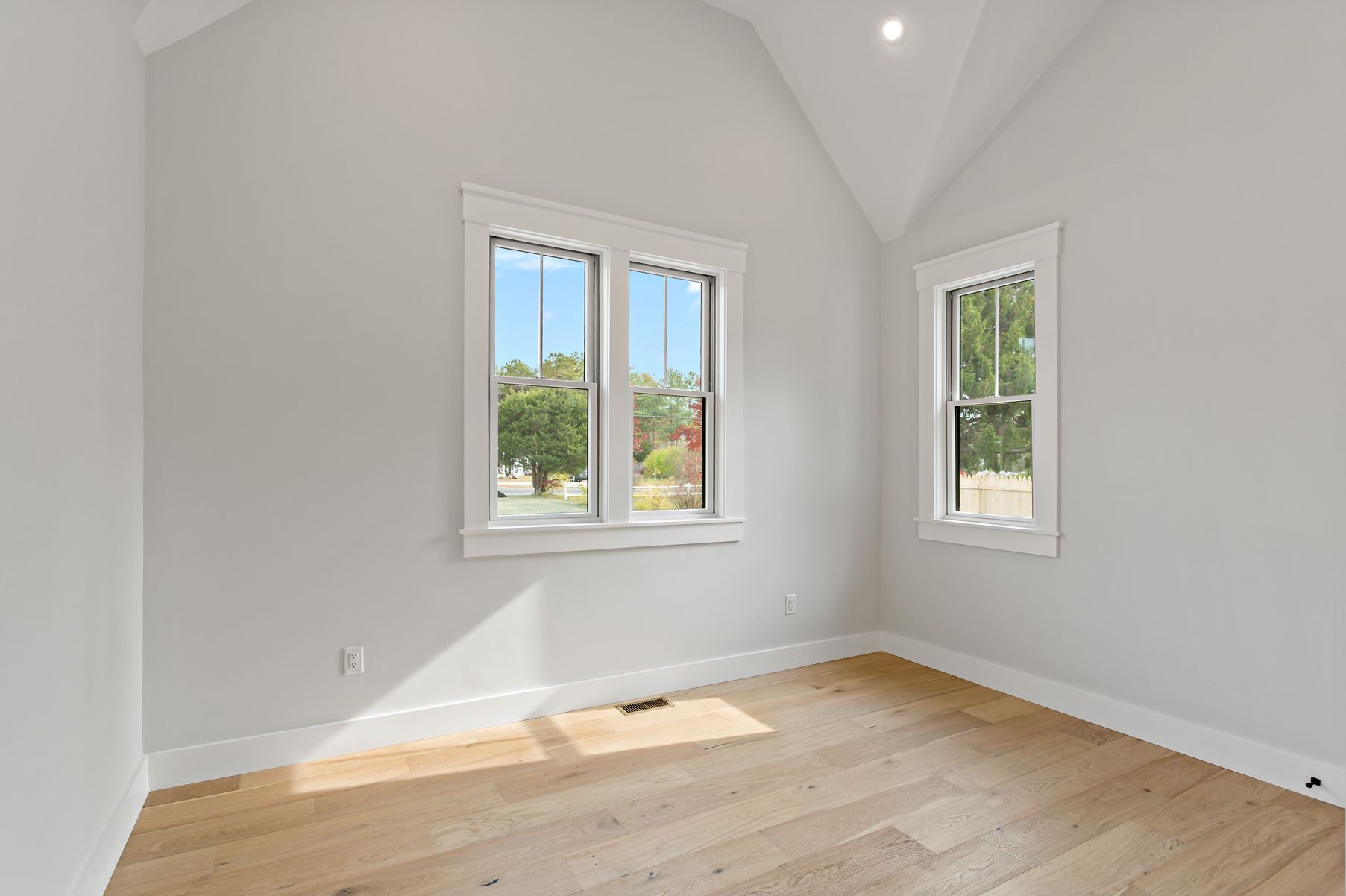 Empty room with light wood floors, white walls, two windows, and vaulted ceiling.