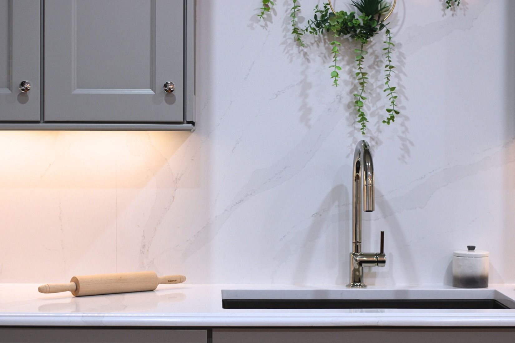 Kitchen sink with faucet, white countertop, gray cabinets, rolling pin, and hanging plant.