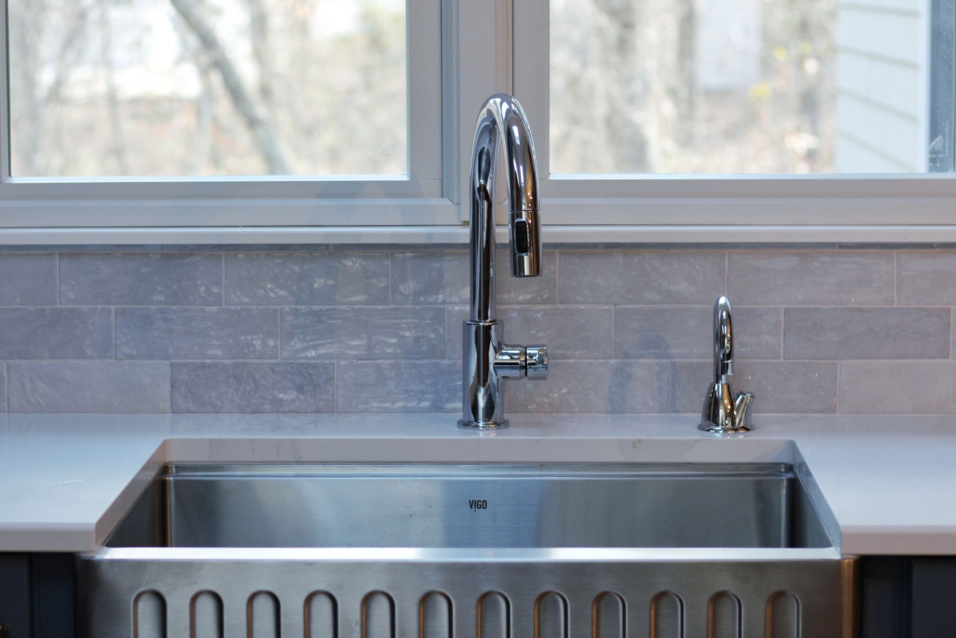 Stainless steel kitchen sink with faucet and window in the background, light-colored backsplash, countertops.