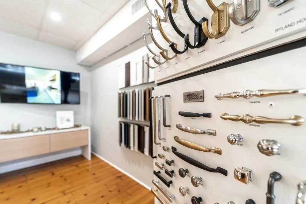 Hardware store display: cabinet pulls, handles, and hooks on a white wall, wooden floor, TV screen.