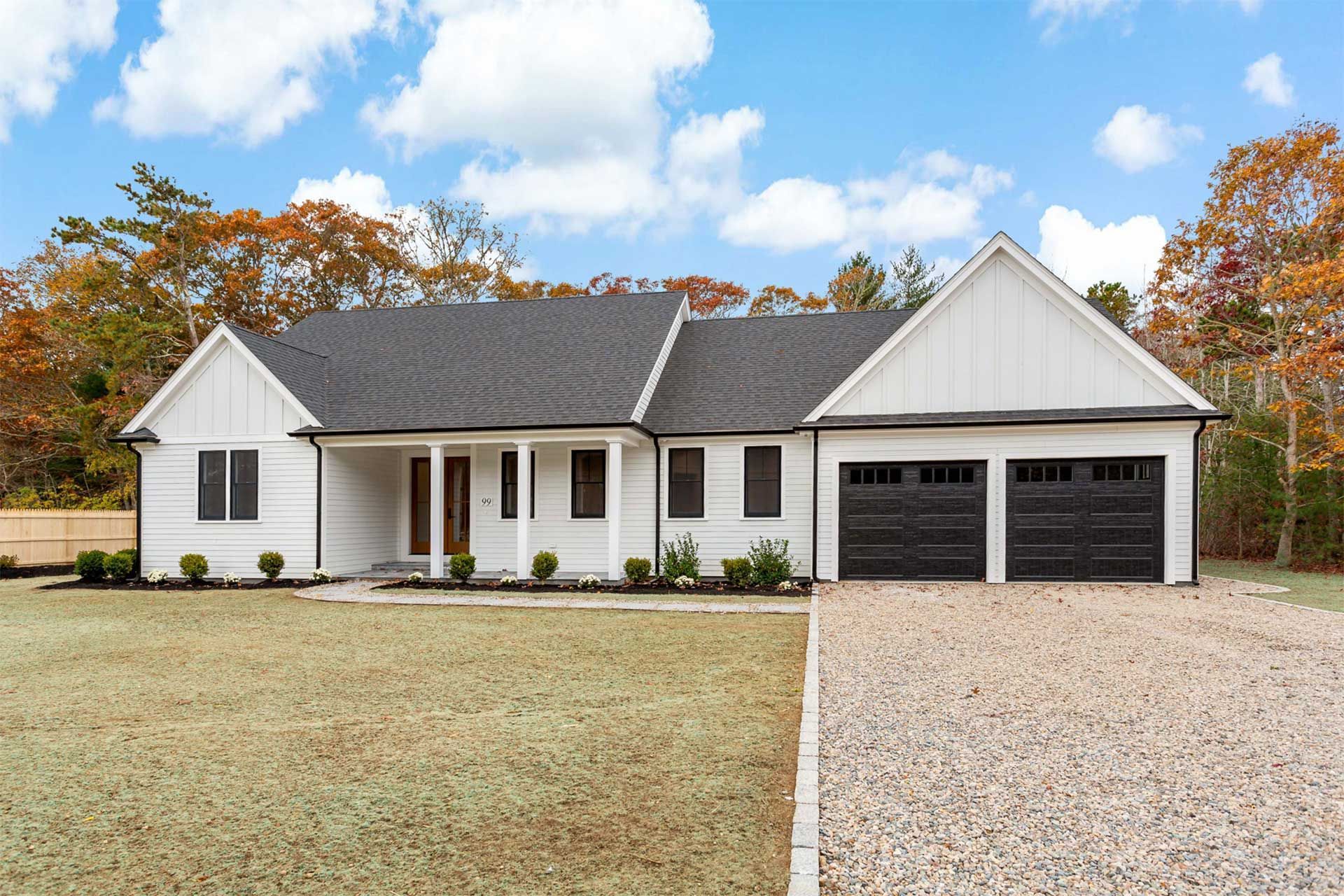 White modern farmhouse with black garage doors, gravel driveway, and autumn trees.