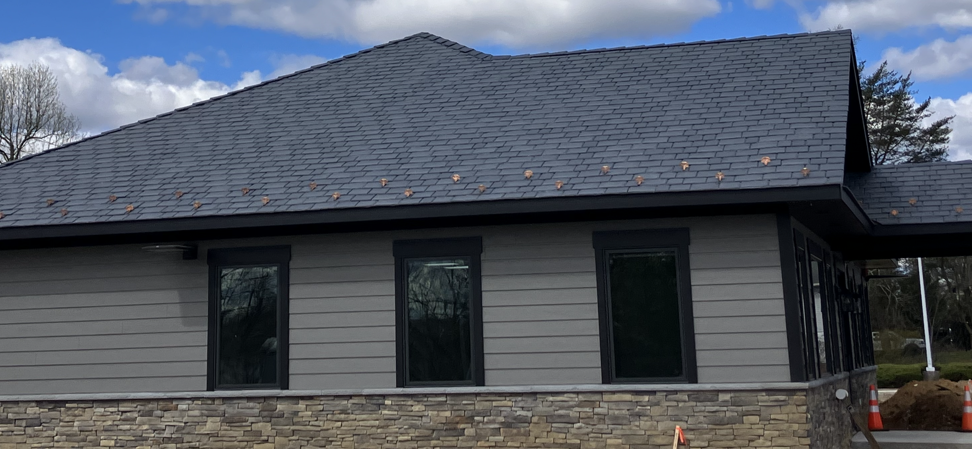 A modern, single-story building with gray lap siding, dark trim, a stone foundation, and a dark shingled roof under a sky.