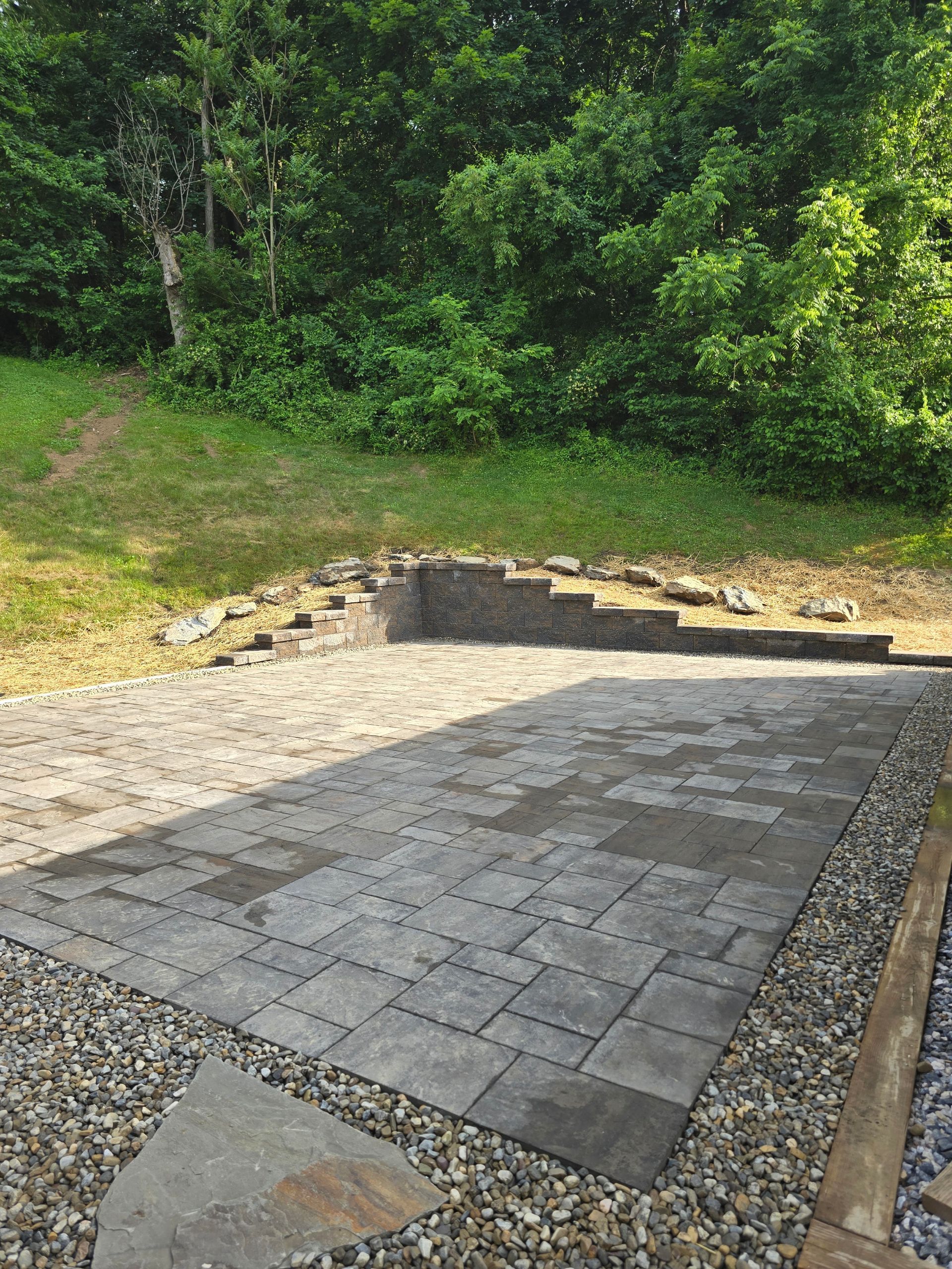 Stone patio under construction beside a grassy slope and dense green trees