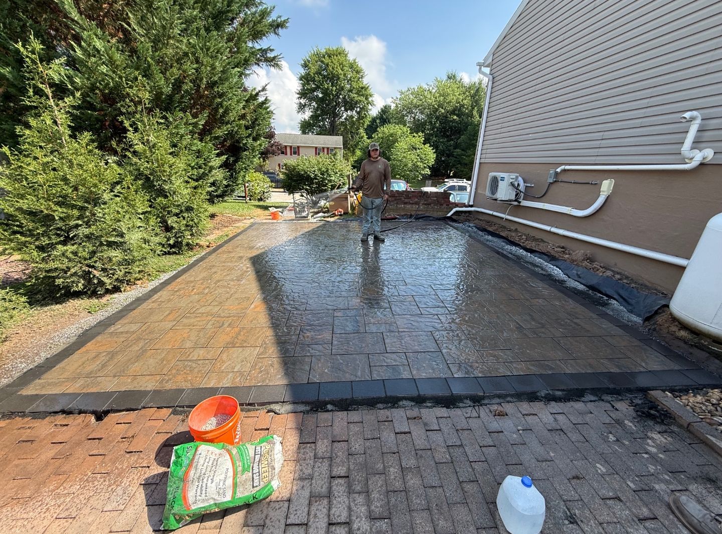 Worker spreading wet concrete on a backyard patio beside a house and garden.