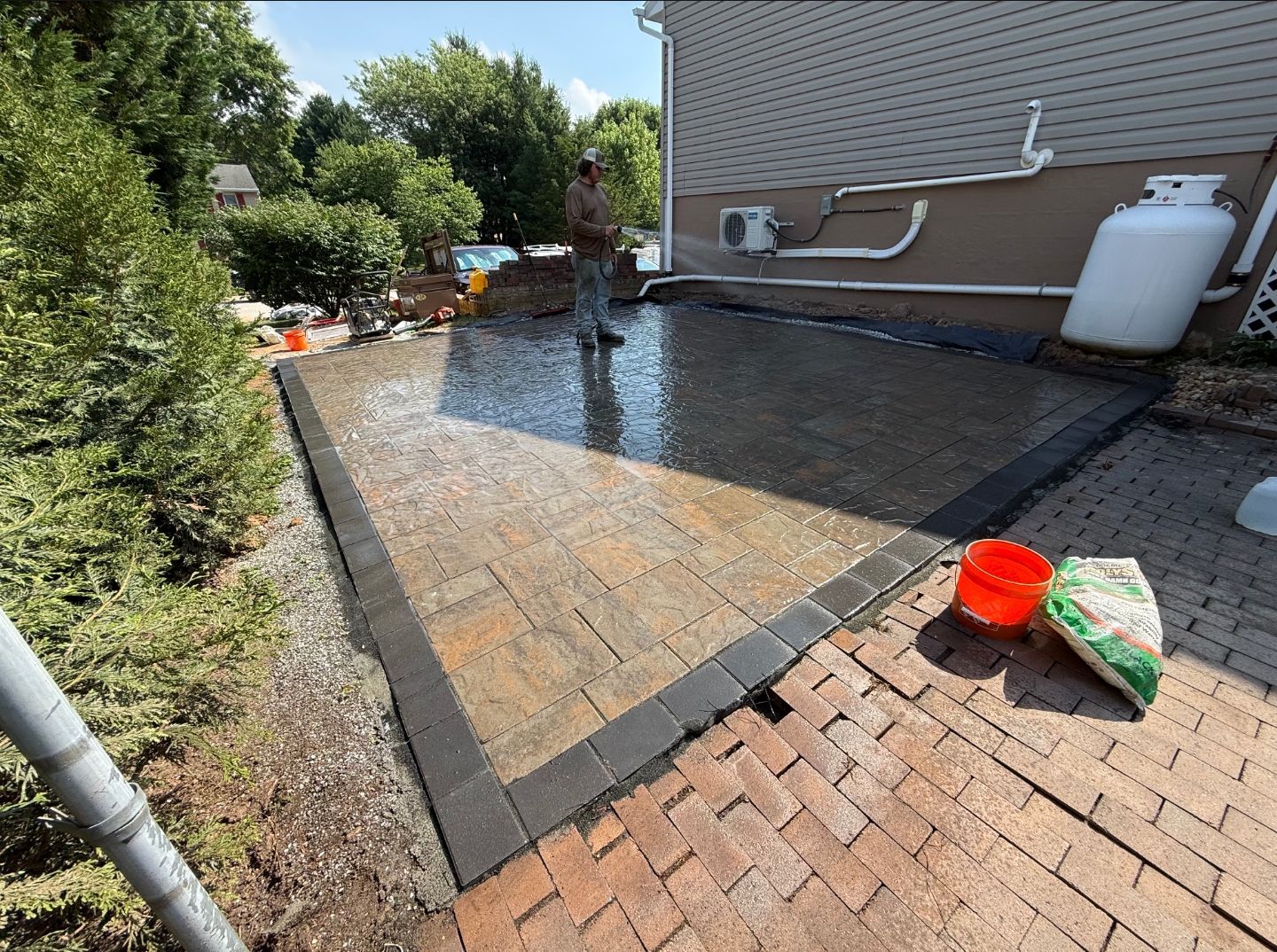 Backyard patio with wet concrete, hose, and red bucket near a brick walkway