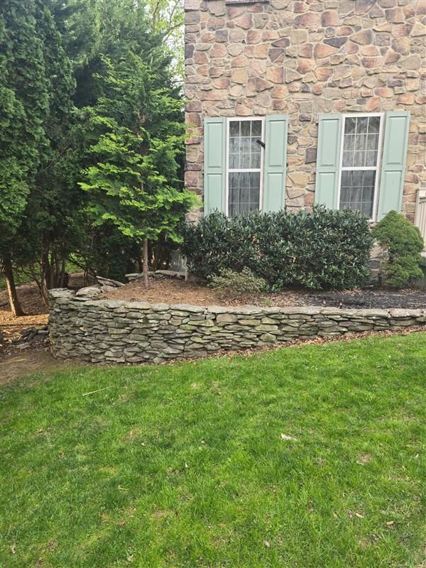 A stone wall is in front of a stone house with blue shutters.