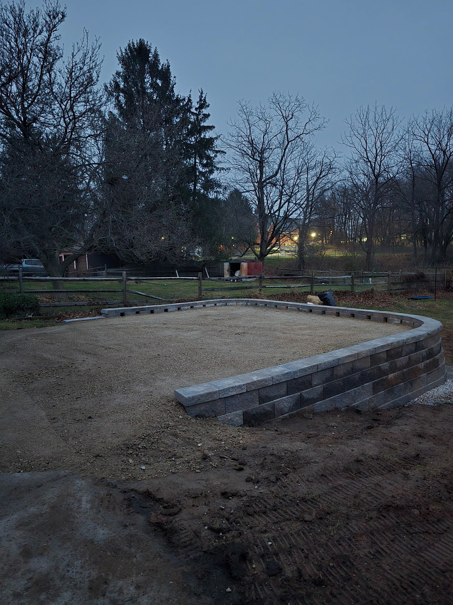 A curved retaining wall made of gray blocks surrounds a gravel area, in a backyard setting.
