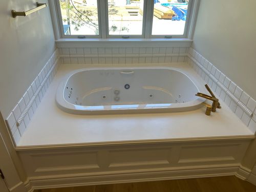 White bathroom with a built-in tub, window above, and gold faucet on a tiled ledge