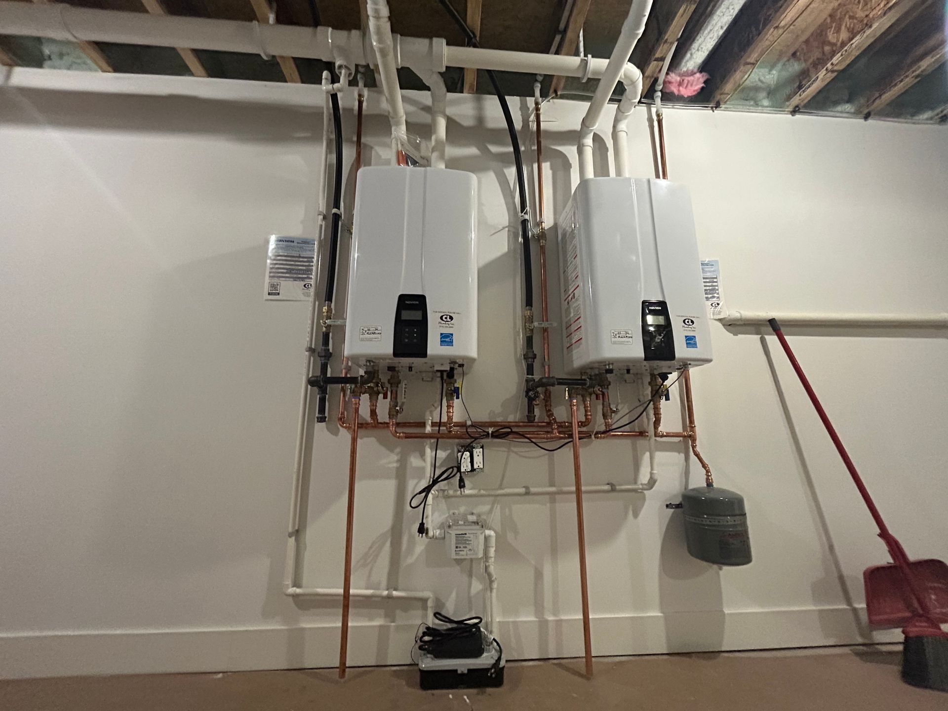 Two white wall-mounted water heaters in a basement with exposed pipes and a black bucket below