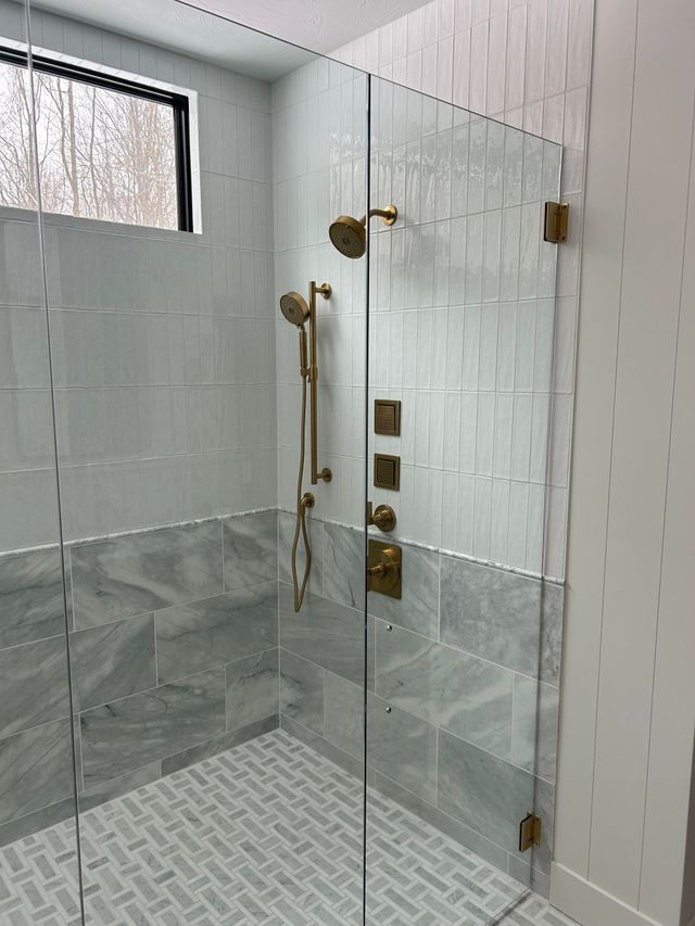 Glass shower enclosure with brass fixtures, white tiled walls, and gray tile floor