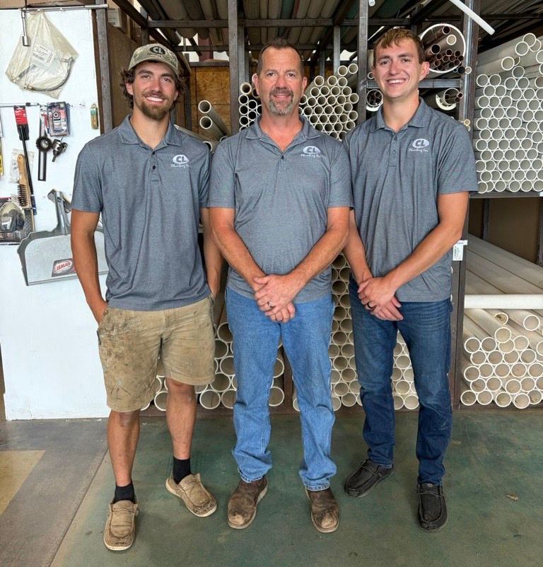 Three men stand in a workshop with shelves and tools behind them, smiling at the camera.