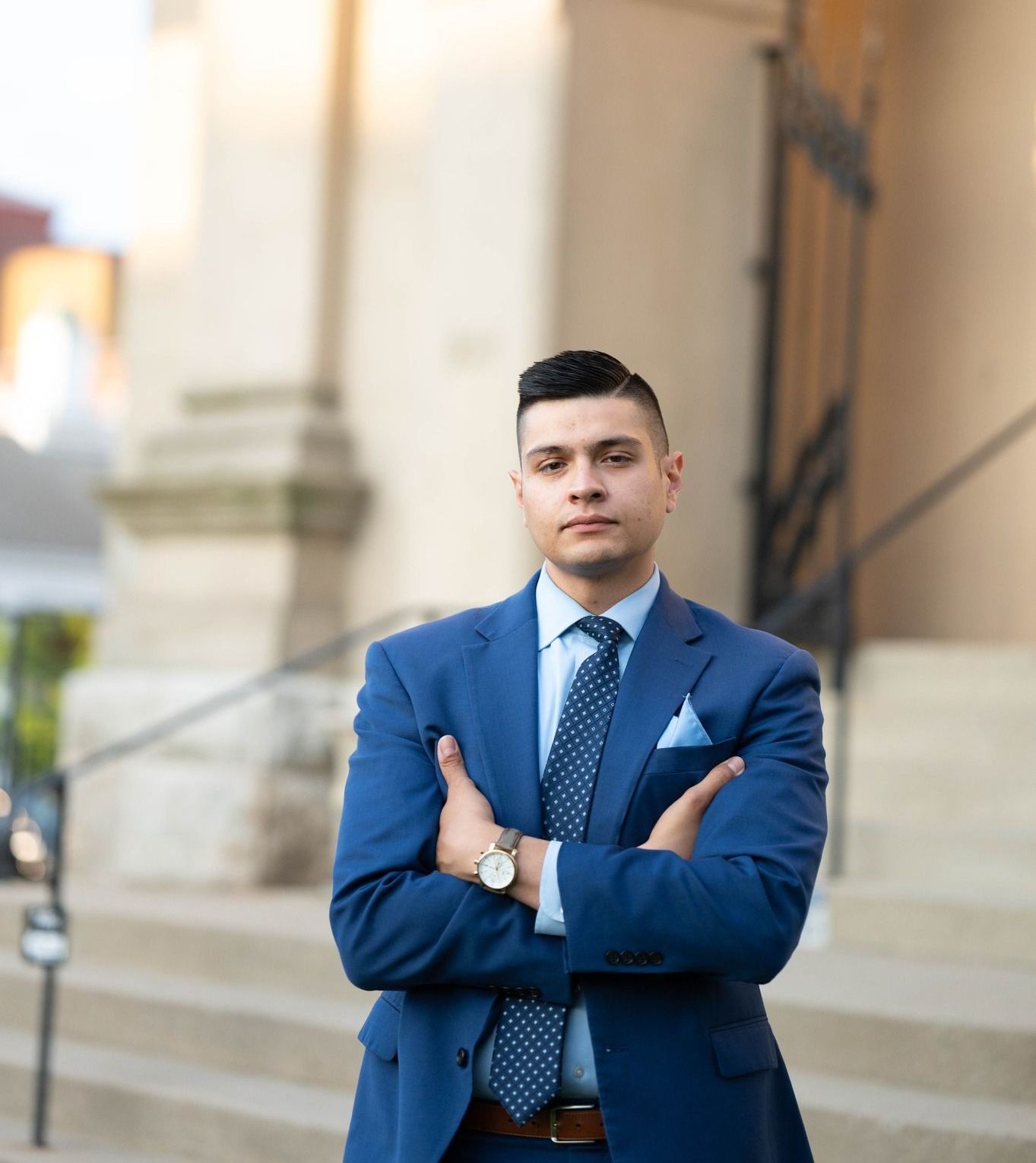 A person in a blue suit and patterned tie stands with arms crossed on stone steps outside a building.