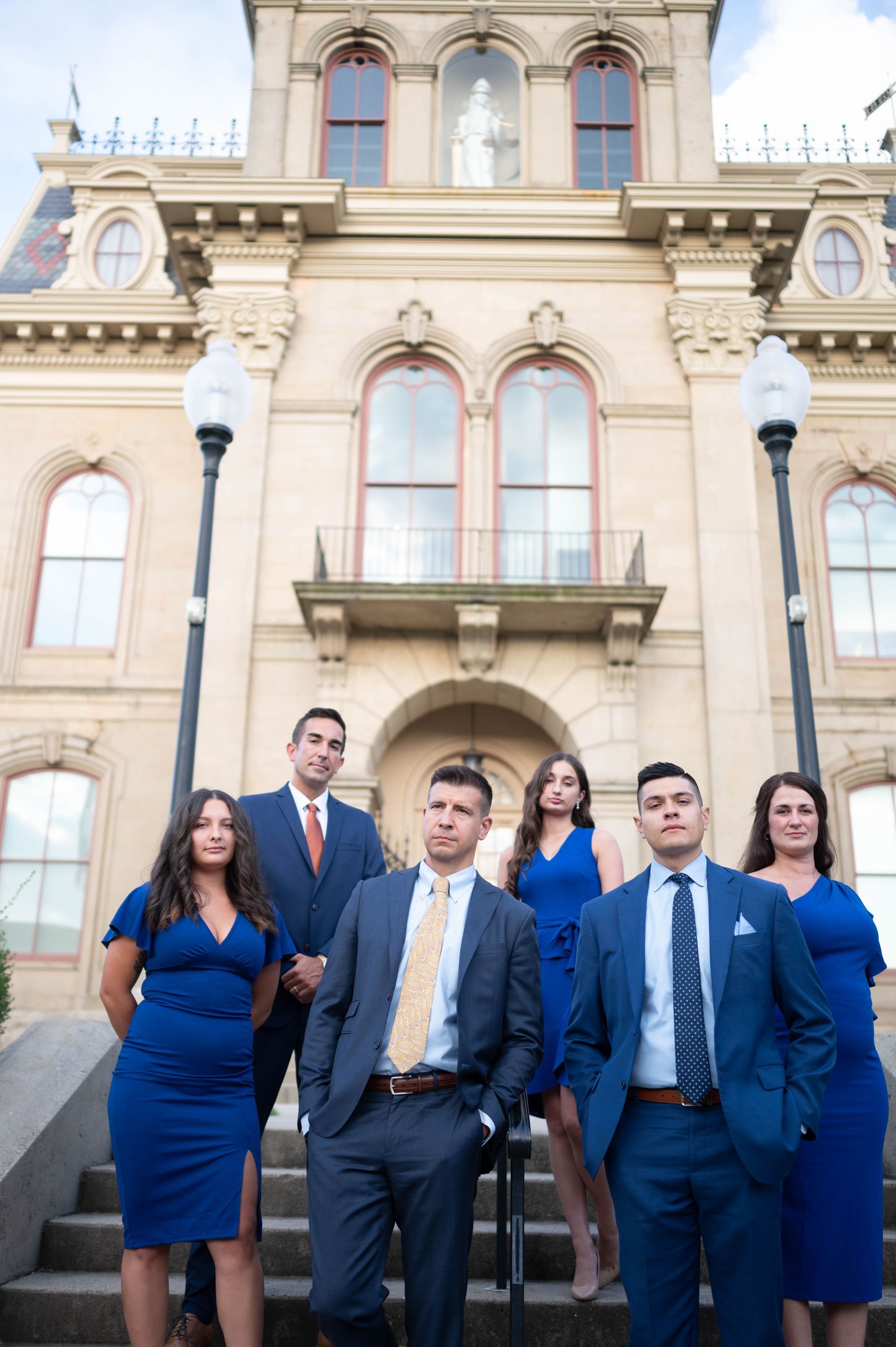 Six people in formal attire stand on stone steps in front of a historic building with large arched windows.