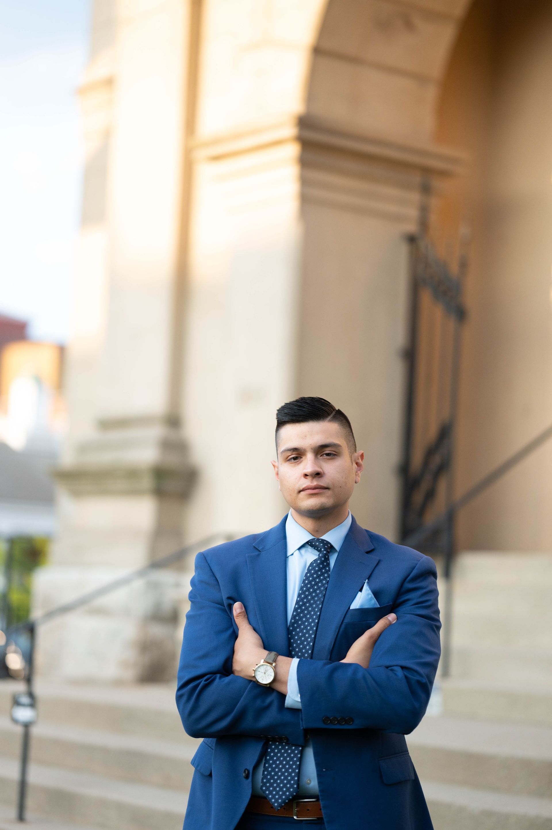 A person in a blue suit and patterned tie stands with arms crossed in front of a stone building with an arched entryway.
