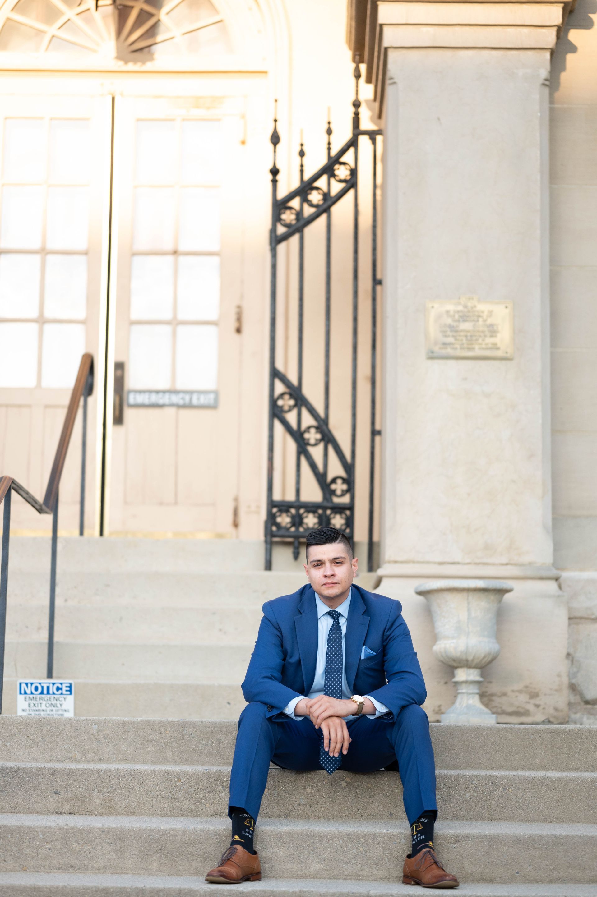 A person in a blue suit and patterned tie sits on stone steps in front of a building with an emergency exit door.