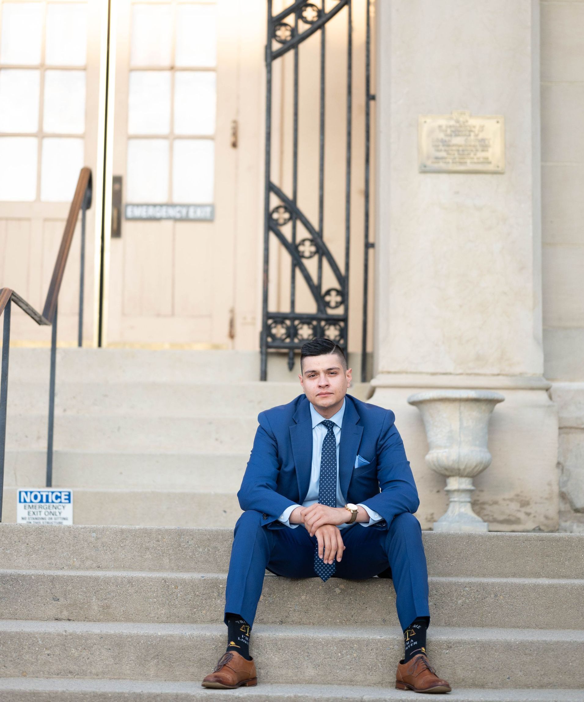 A person in a blue suit and patterned socks sits on stone steps in front of a building entrance with a metal gate.