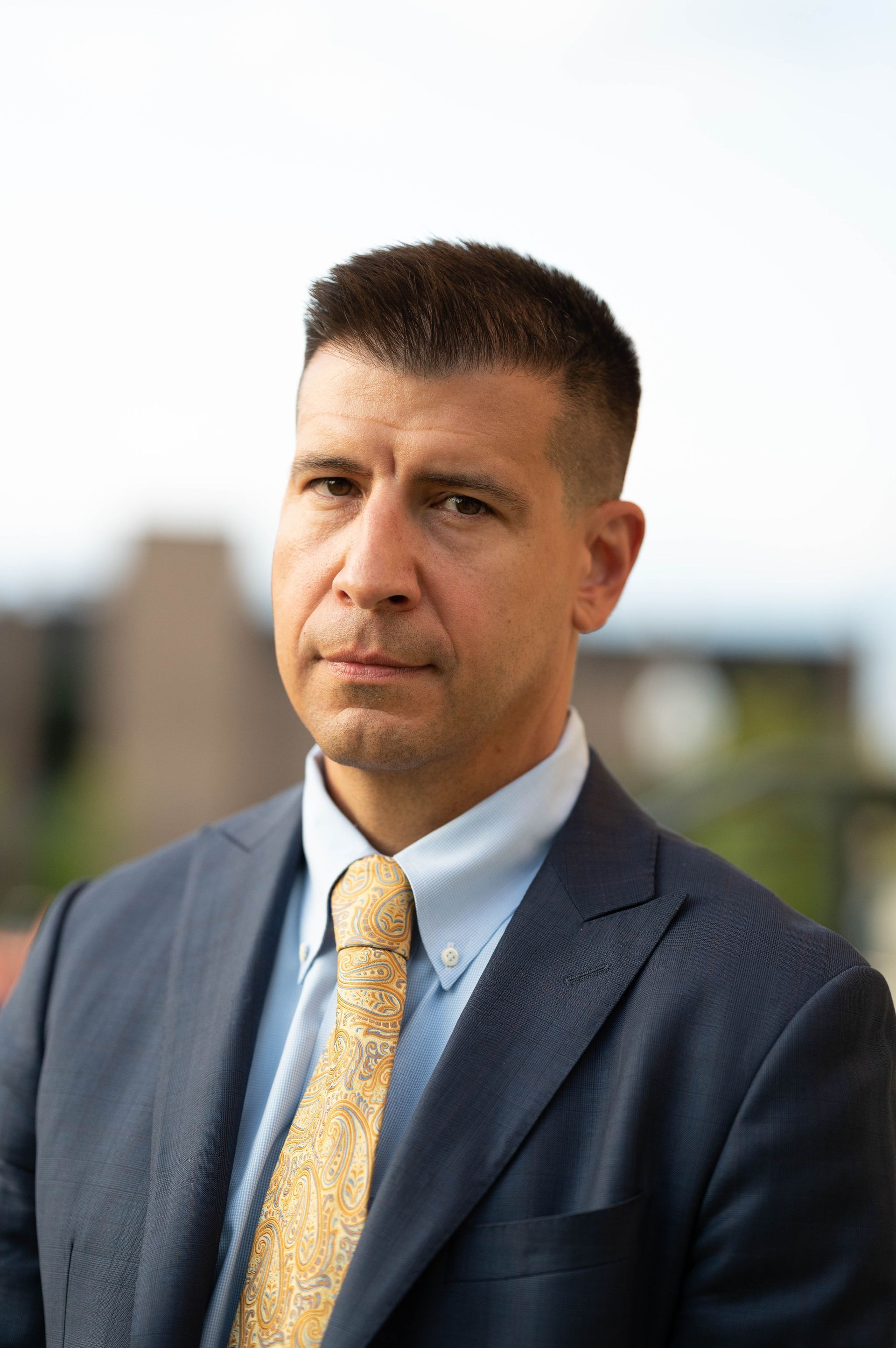 A person with short, dark hair wears a navy blue suit, light blue shirt, and gold-patterned tie, posing outdoors.