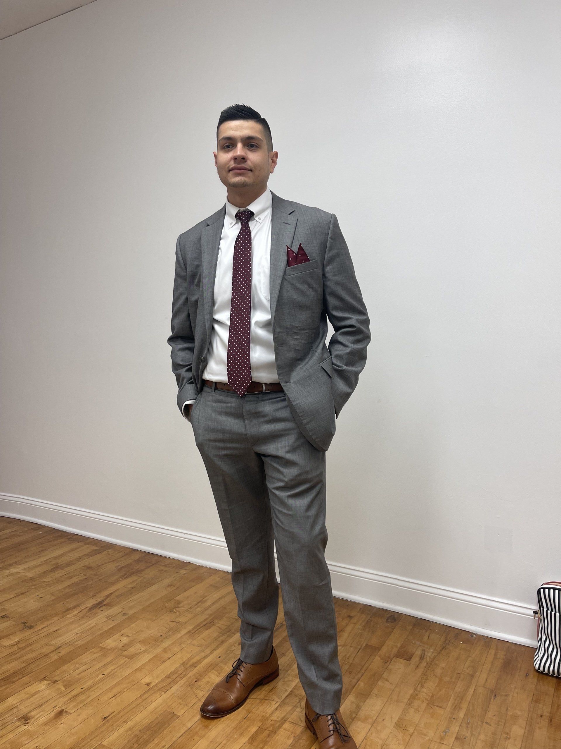 A person in a grey suit, white shirt, and maroon tie stands with hands in pockets against a plain white wall.
