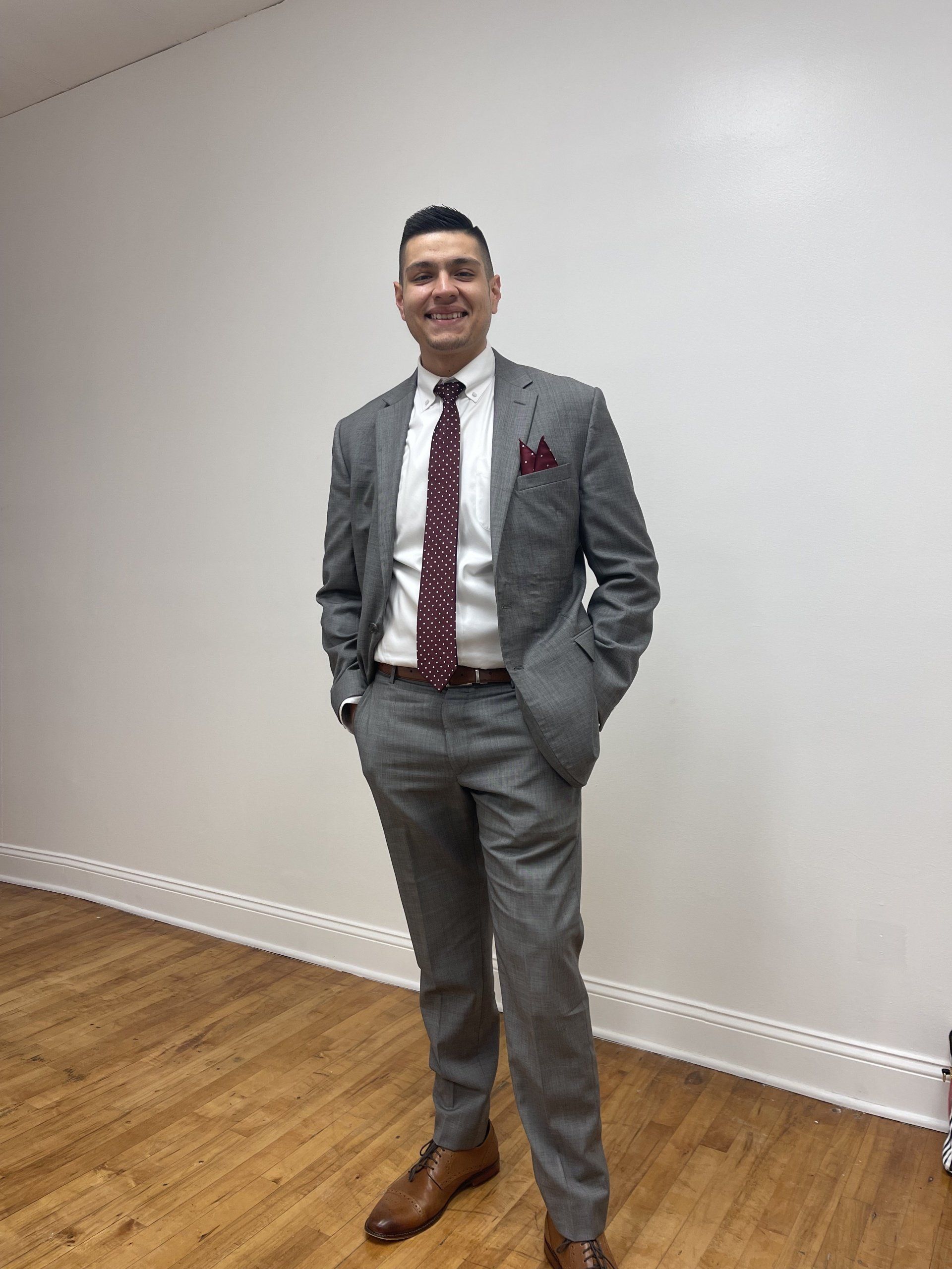 A smiling person wearing a grey suit, white shirt, and maroon patterned tie, standing in a room with white walls.