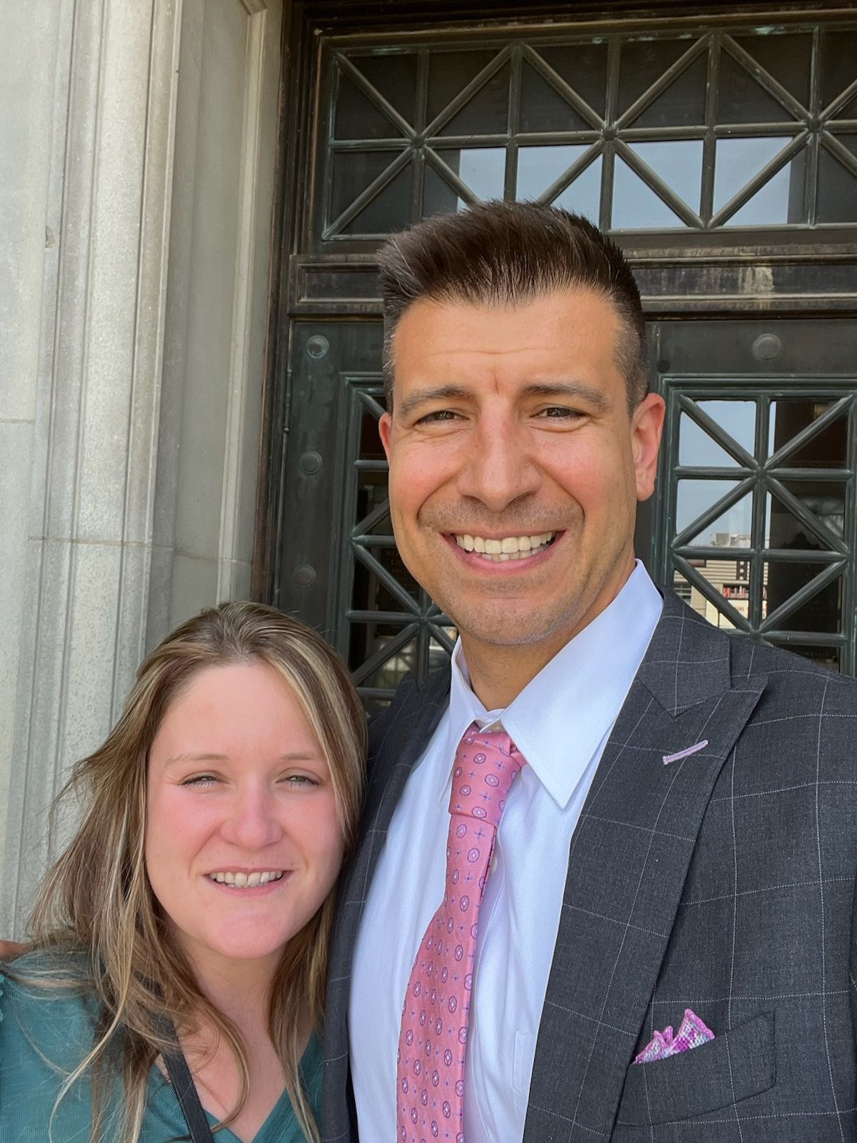 A woman and a man in a suit and pink tie stand side-by-side in front of a dark wooden door with a glass grid design.