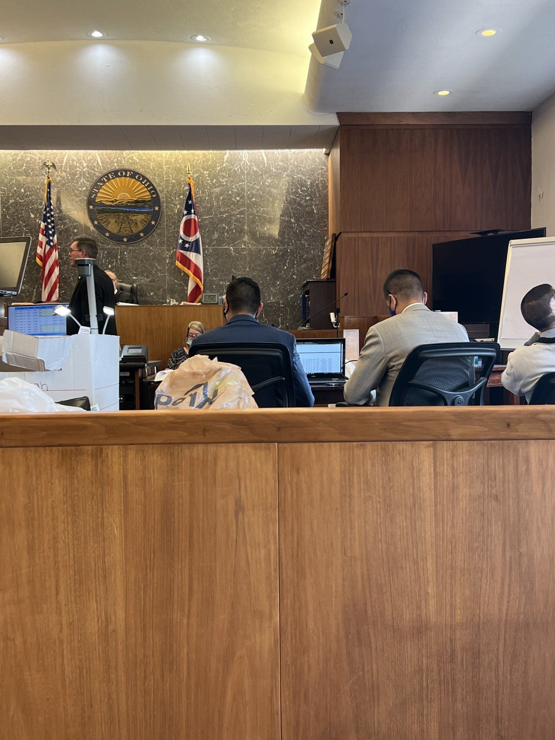 People sit at desks in a wood-paneled courtroom, with the American and Ohio state flags displayed on the back wall.