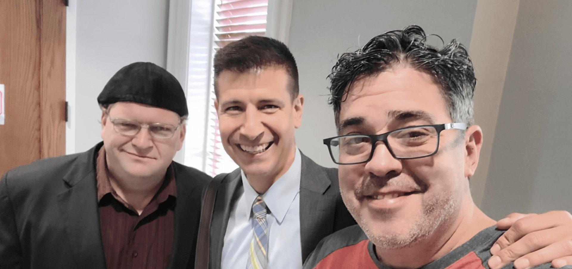 Three people smiling together for a selfie indoors, wearing casual and formal attire.