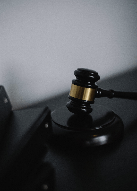 A black wooden judge's gavel with a gold band rests on a matching sounding block against a plain grey background.