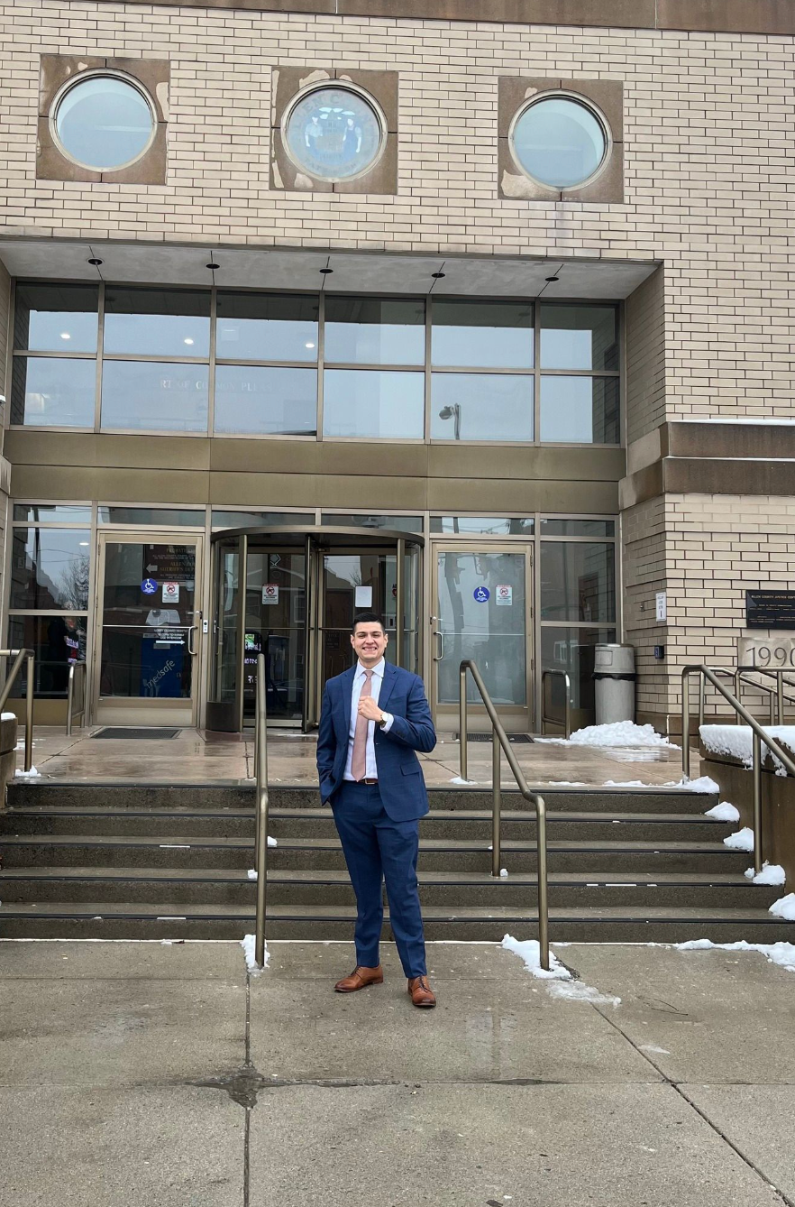 A person in a blue suit and light tie stands on concrete steps in front of a building entrance with circular windows.