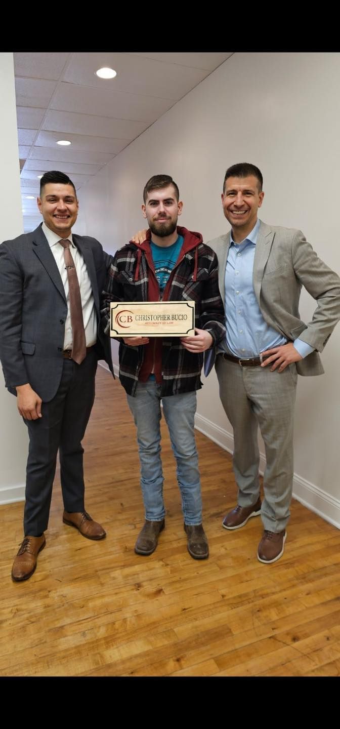 Three men standing in a hallway, the center person holding a wooden award plaque, smiling at the camera.
