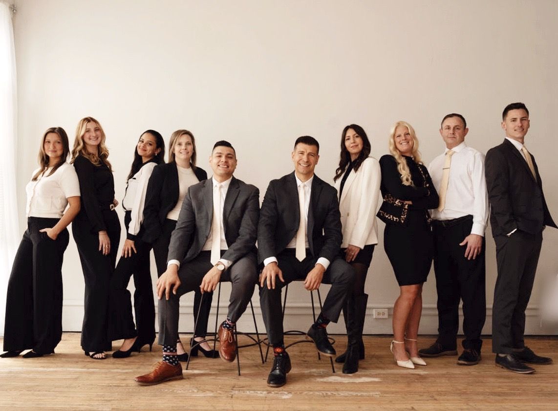 A group of ten professionals in formal business attire pose for a studio portrait against a plain, light-colored wall.