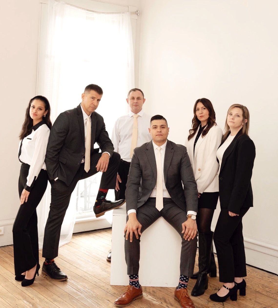 Six people in formal business attire pose for a group portrait in a bright, minimalist room with wooden floors.