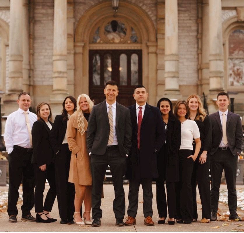 Group of ten professional colleagues posing in suits and business attire in front of a stone building with columns.