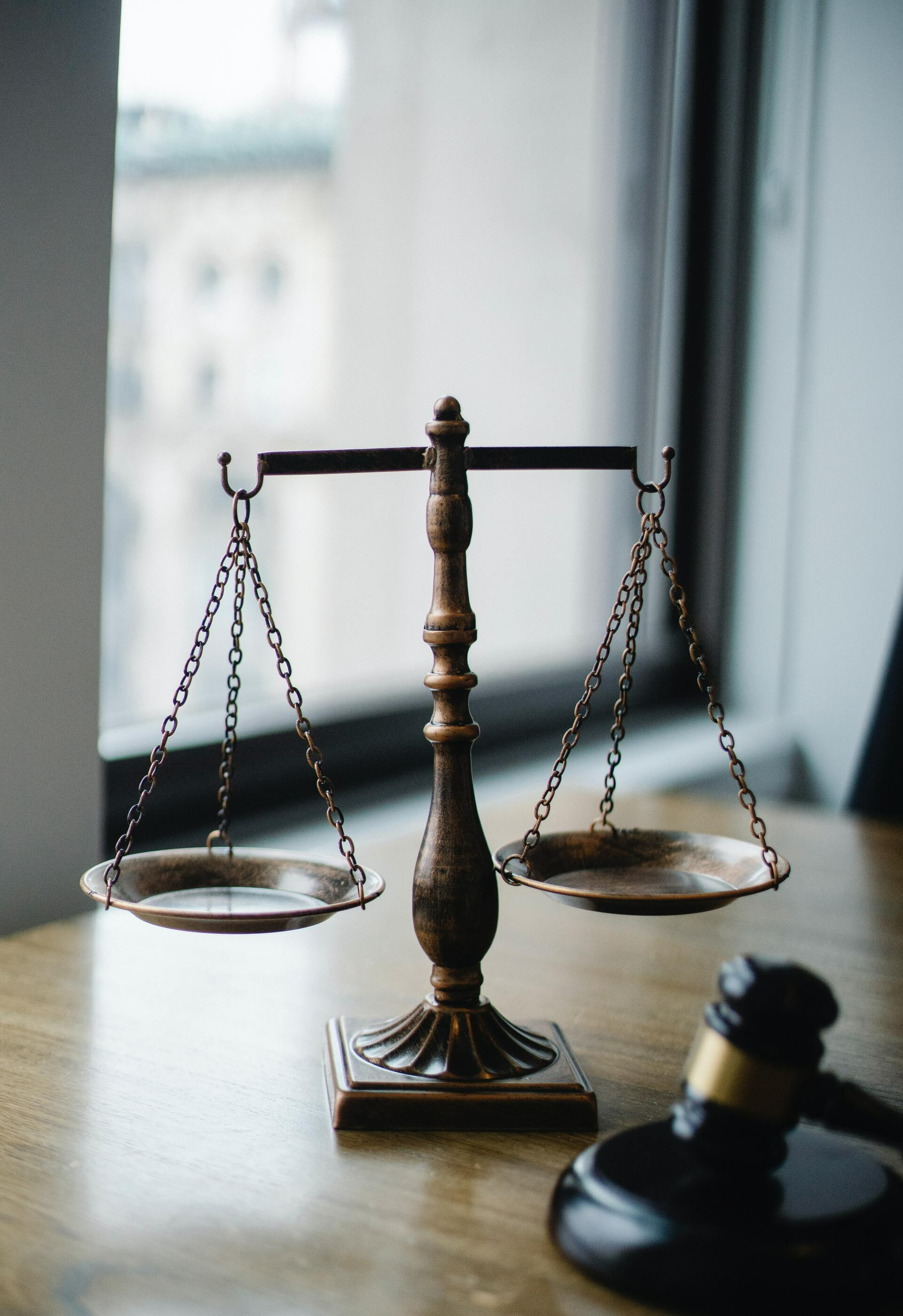 A vintage brass balance scale sits on a wooden table next to a judge’s gavel in front of a window.