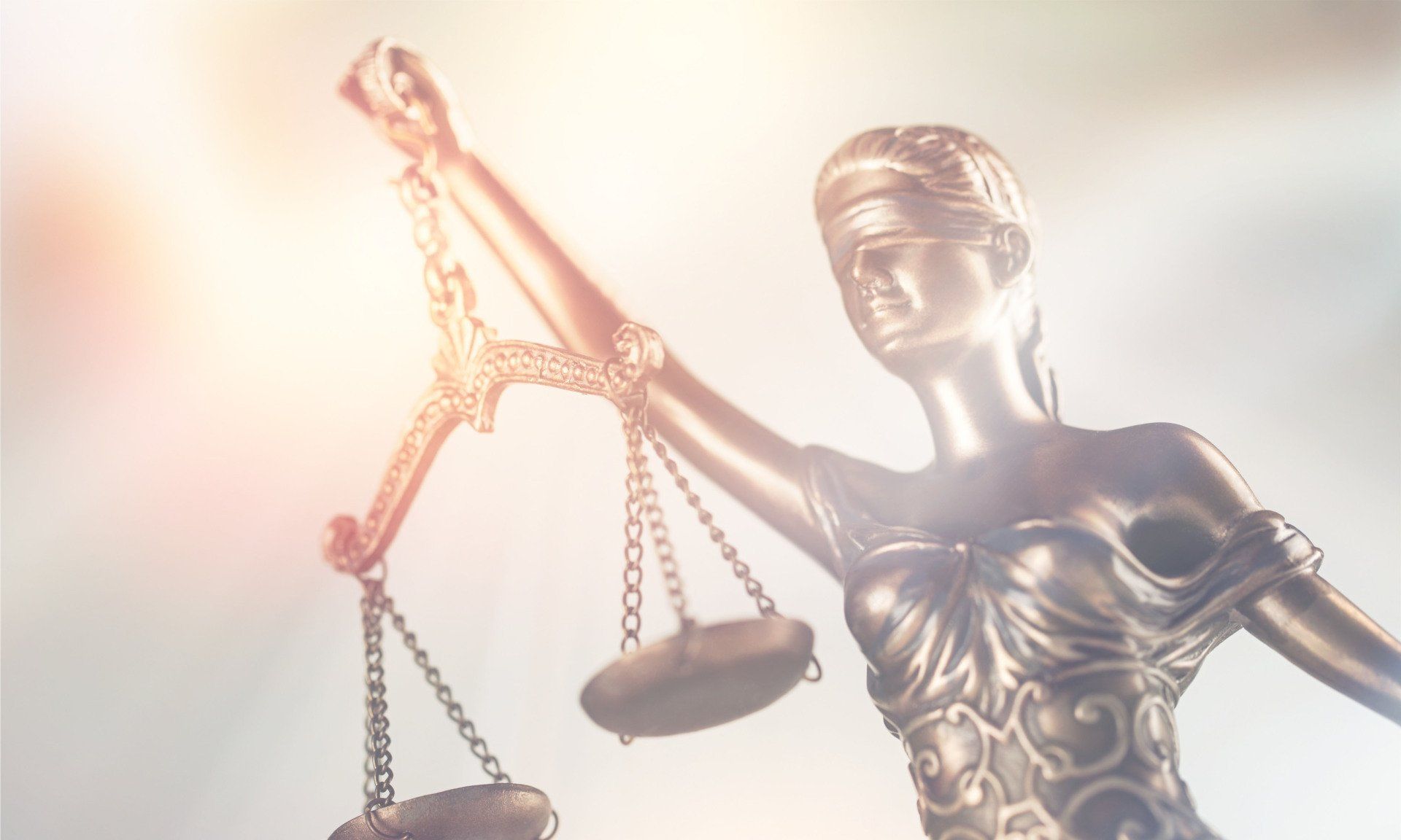 A statue of Lady Justice holding scales, illuminated by a warm, glowing light against a bright, hazy background.