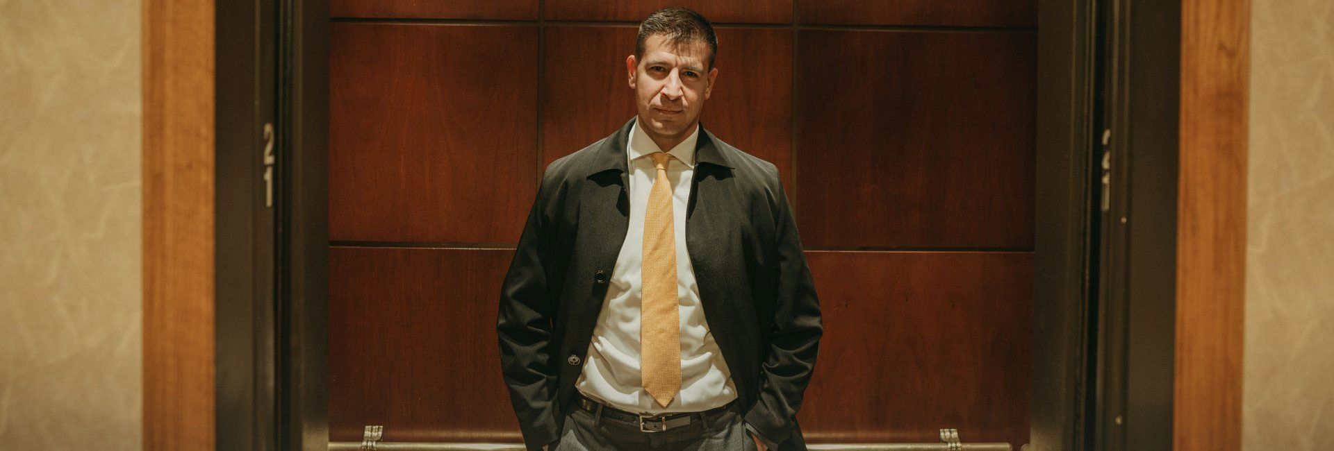 A person in a black jacket and yellow tie stands with hands in pockets in front of a dark wood-paneled door.