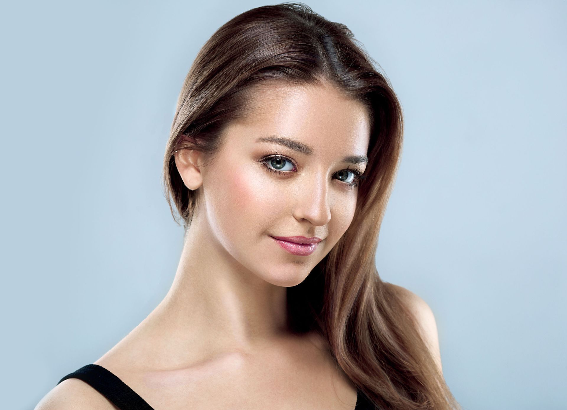 Woman with long brown hair, smiling at the viewer, wearing a black top against a light blue background.