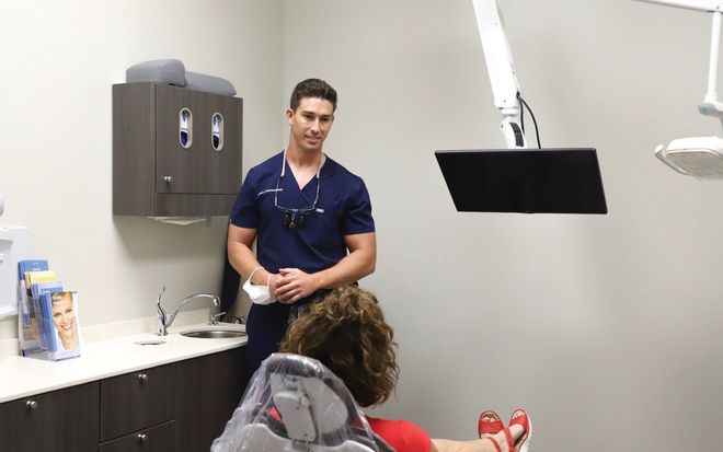 Dentist in blue scrubs stands over a patient in a dental chair in a modern office.