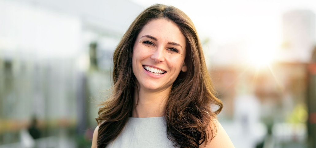 Woman with long brown hair smiles at the camera in front of a blurred outdoor background.