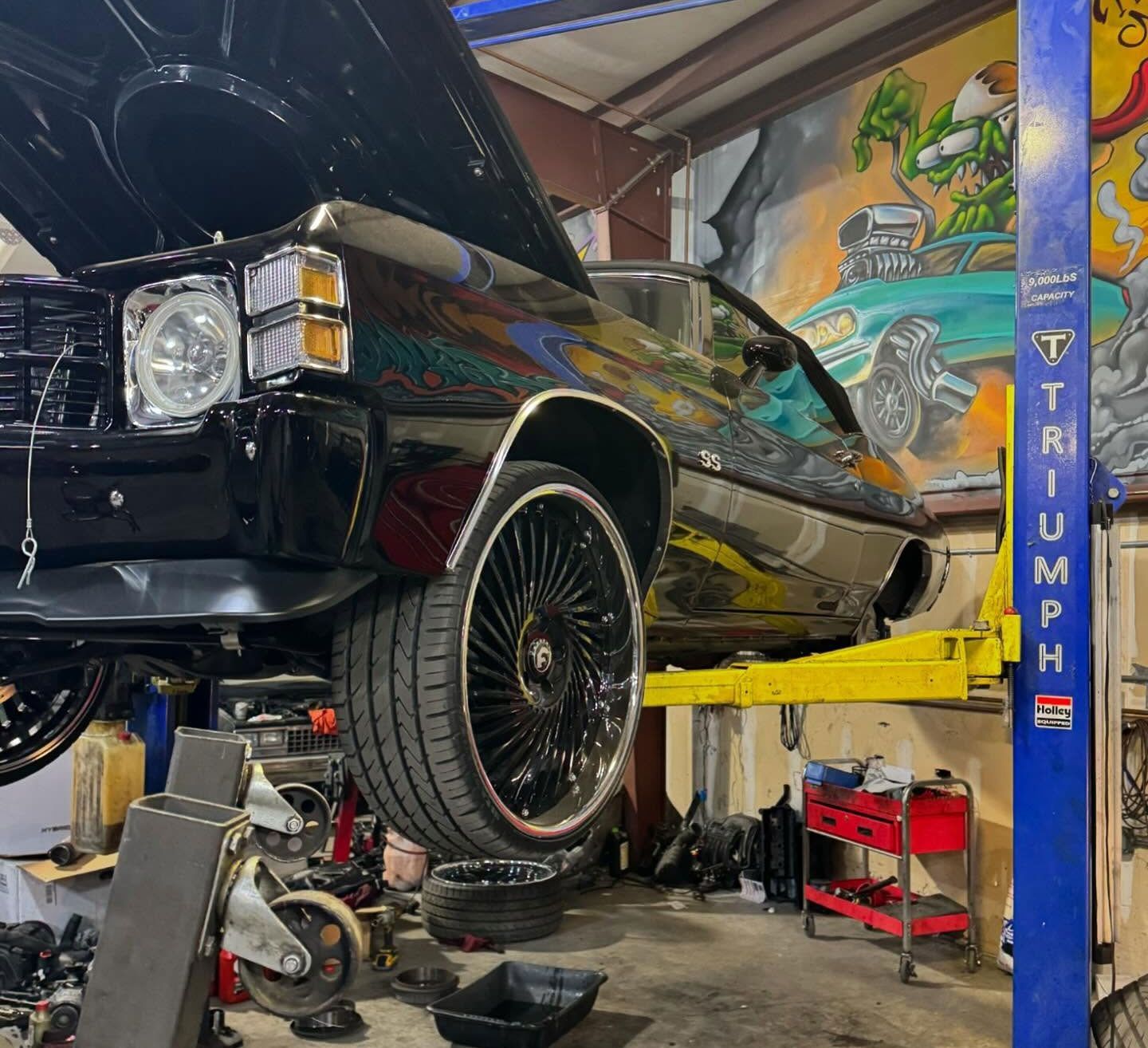 A black classic Chevrolet Chevelle SS raised on a yellow auto lift in a garage with a mural on the back wall.