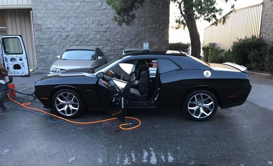 A black Dodge Challenger parked outdoors with its driver-side door open and an orange extension cord nearby.