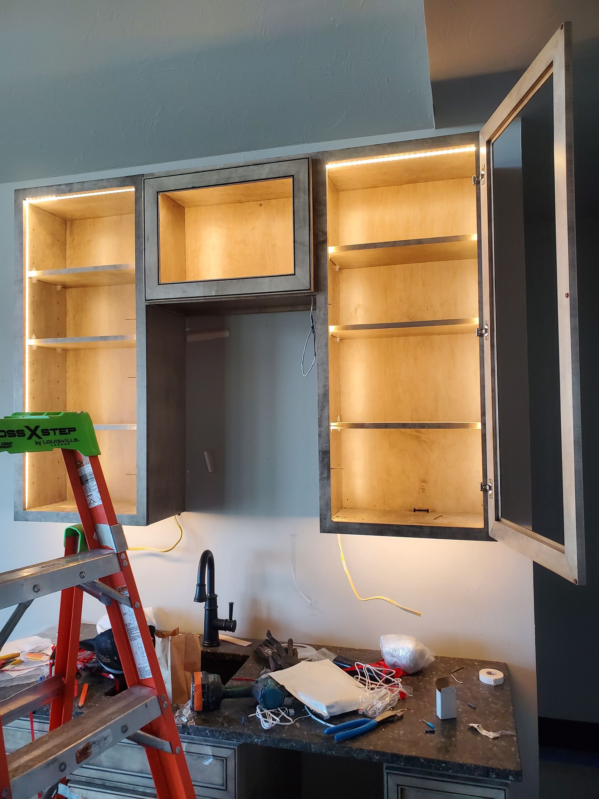 Kitchen cabinets with interior lighting are installed on a wall. A ladder and countertop are in front.